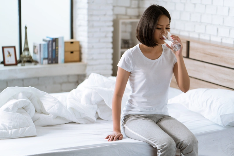 asian women drinking water on edge of bed