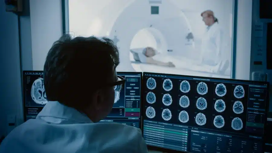 A doctor looking at brain scans on a computer screen while there is a woman with Dry Mouth a Symptom of Covid in an MRI machine in the background