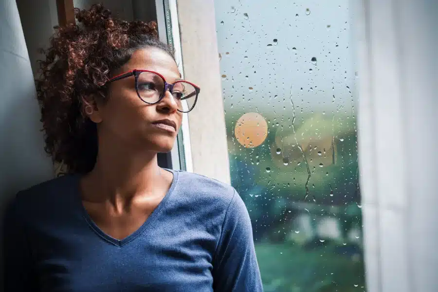 A woman with glasses looking out a window on a rainy day depressed because she has Dry Mouth a Symptom of Covid