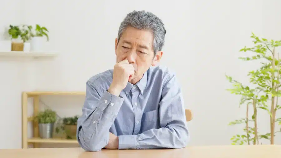 An elderly man with Dry Mouth a Symptom of Covid sitting at a table looking contemplative with his elbow rested on the table and his hand to his chin